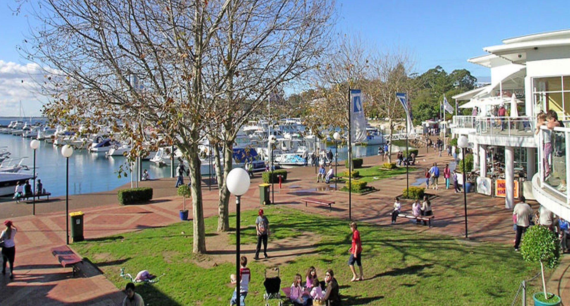 A marina with boats, a park area with benches and people, and a building with a balcony.