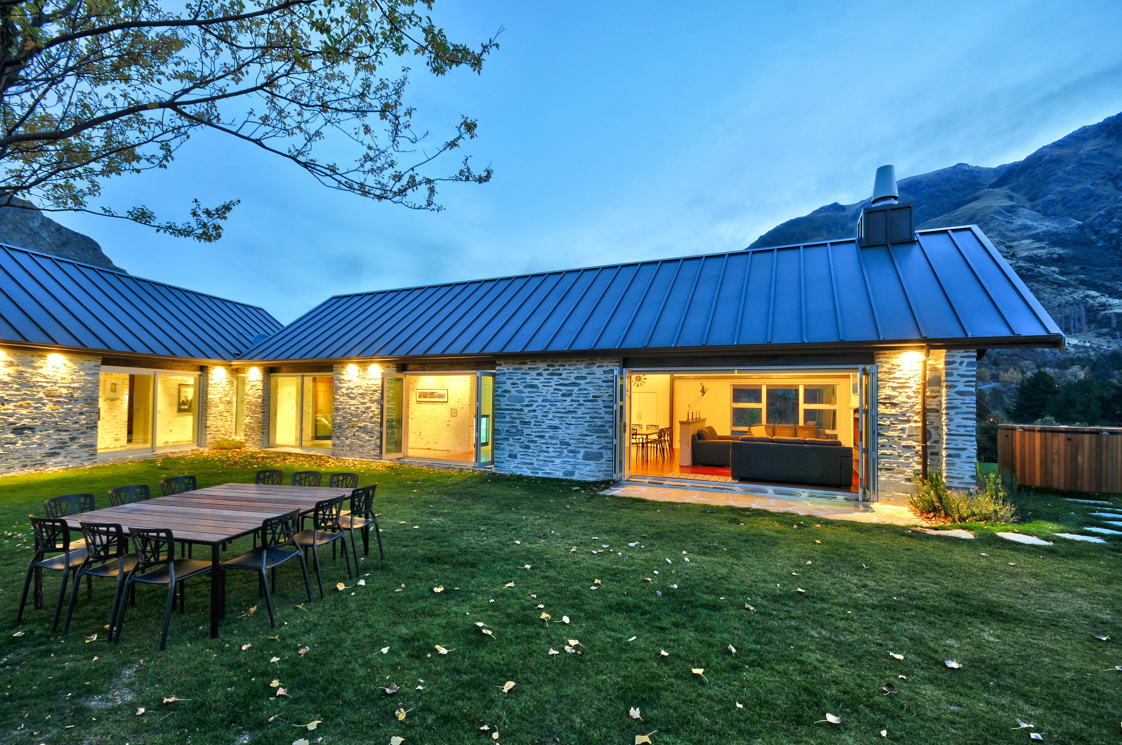 A modern house with a stone exterior, a large patio with a dining table and chairs, and a view of mountains in the background.