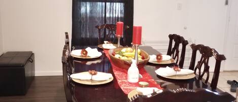 A dining room with a wooden table set for six, surrounded by chairs. There is a chandelier above the table, a framed picture on the wall, and a window with dark curtains.