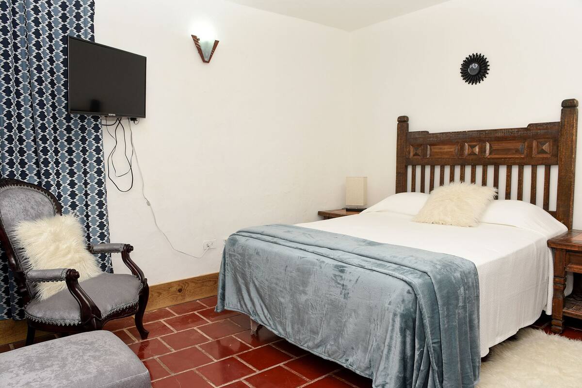 A bedroom with a wooden bed, a chair with a fluffy pillow, a television on the wall, and a patterned curtain.