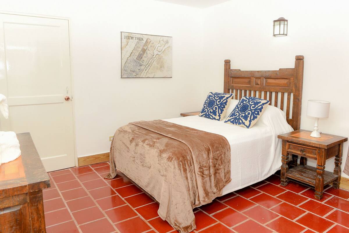 A bedroom with a wooden bed, a nightstand with a lamp, a map on the wall, and a red-tiled floor.