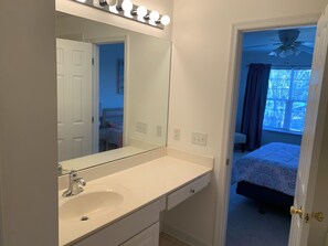 Une salle de bain avec un lavabo blanc, un miroir avec éclairage intégré et une porte donnant sur une chambre à coucher équipée d’un lit et d’une fenêtre.