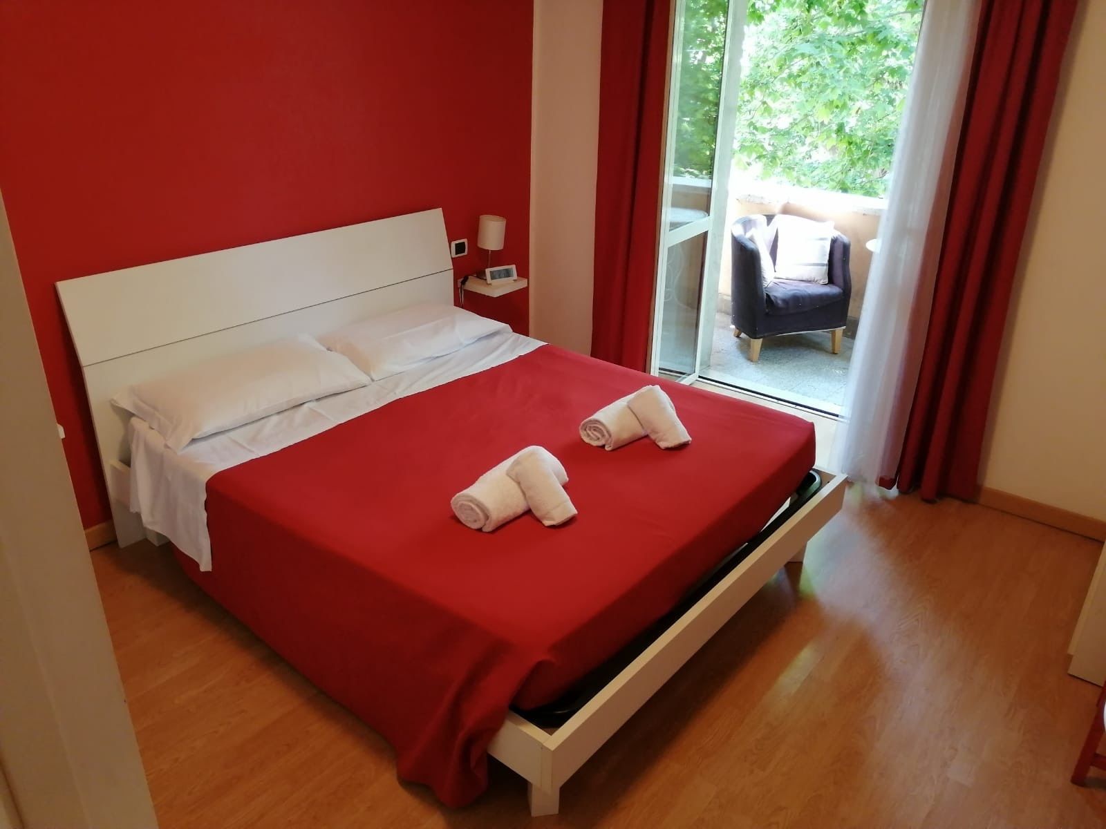 A bed with a red bedspread and two rolled towels, a white headboard, and a small bedside table with a lamp.