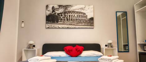 A hotel room with a bed, a heart-shaped pillow, a picture of the Colosseum, a mirror, and a nightstand with a lamp.