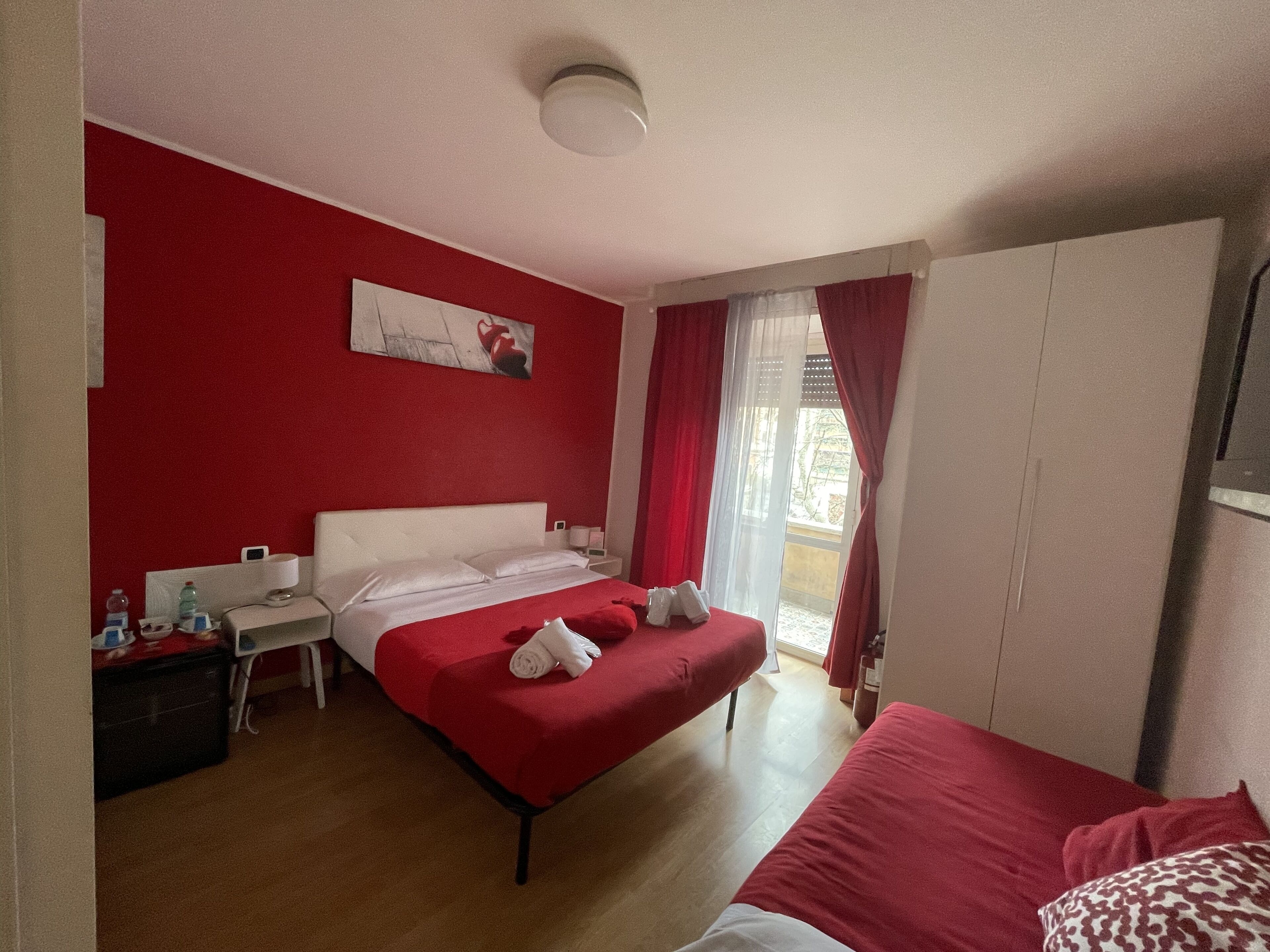 A hotel room with a red and white color scheme, a bed with red bedding, a bedside table with a lamp, and a window with red curtains.