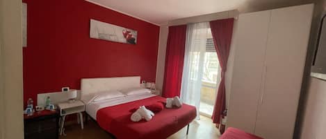 Una habitación de hotel con una decoración en rojo y blanco, una cama con ropa de cama roja, una mesita de noche con una lámpara y una ventana con cortinas rojas.