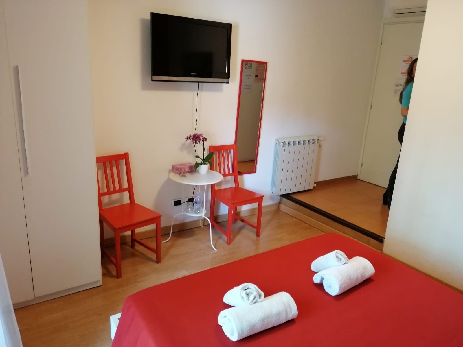 Una camera d'albergo con un letto, due poltrone rosse, un tavolino rotondo con una pianta, uno specchio e una TV a parete.