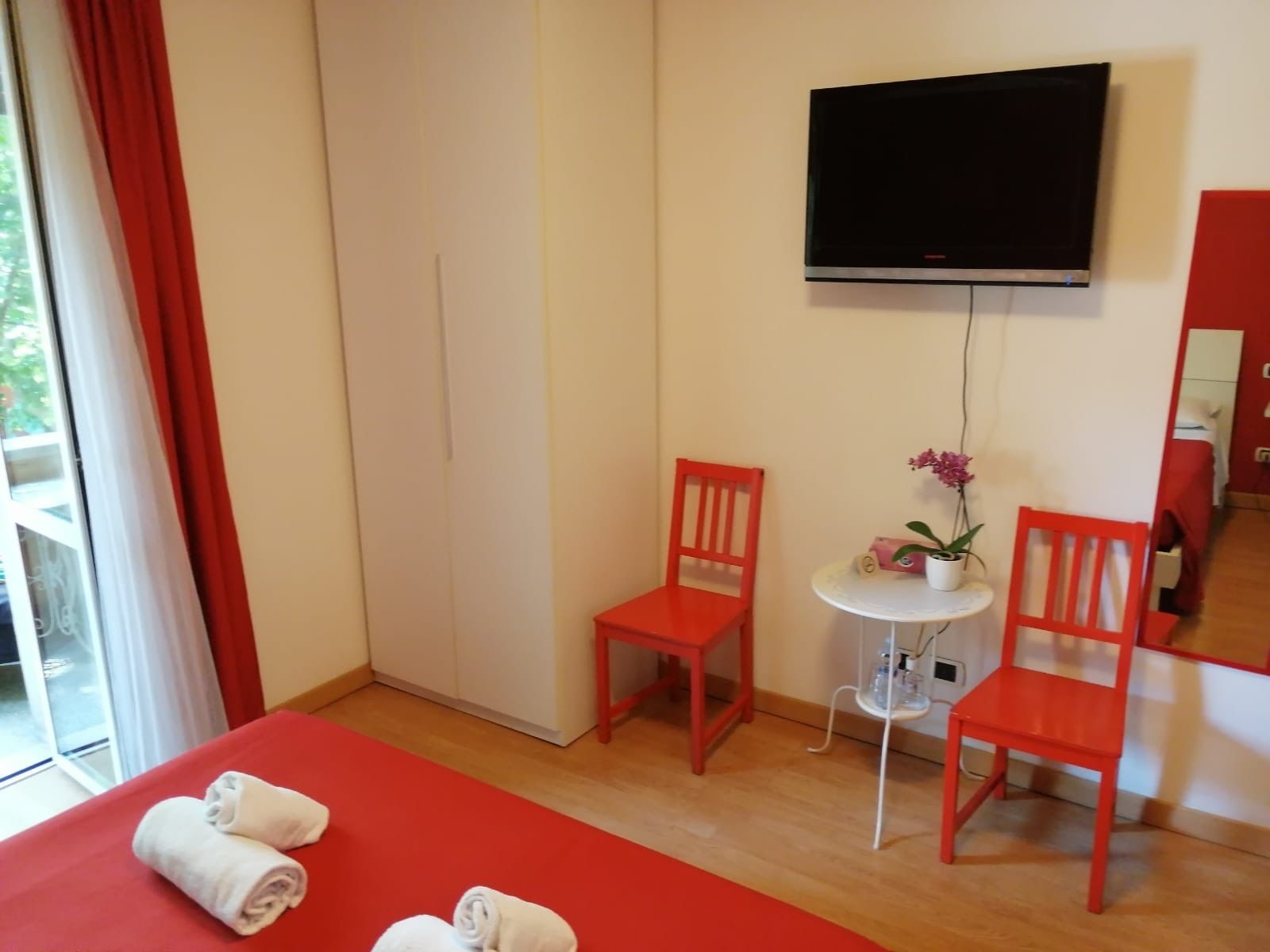 A hotel room with a bed, two red chairs, a small white table with a plant, a TV mounted on the wall, and a wardrobe.