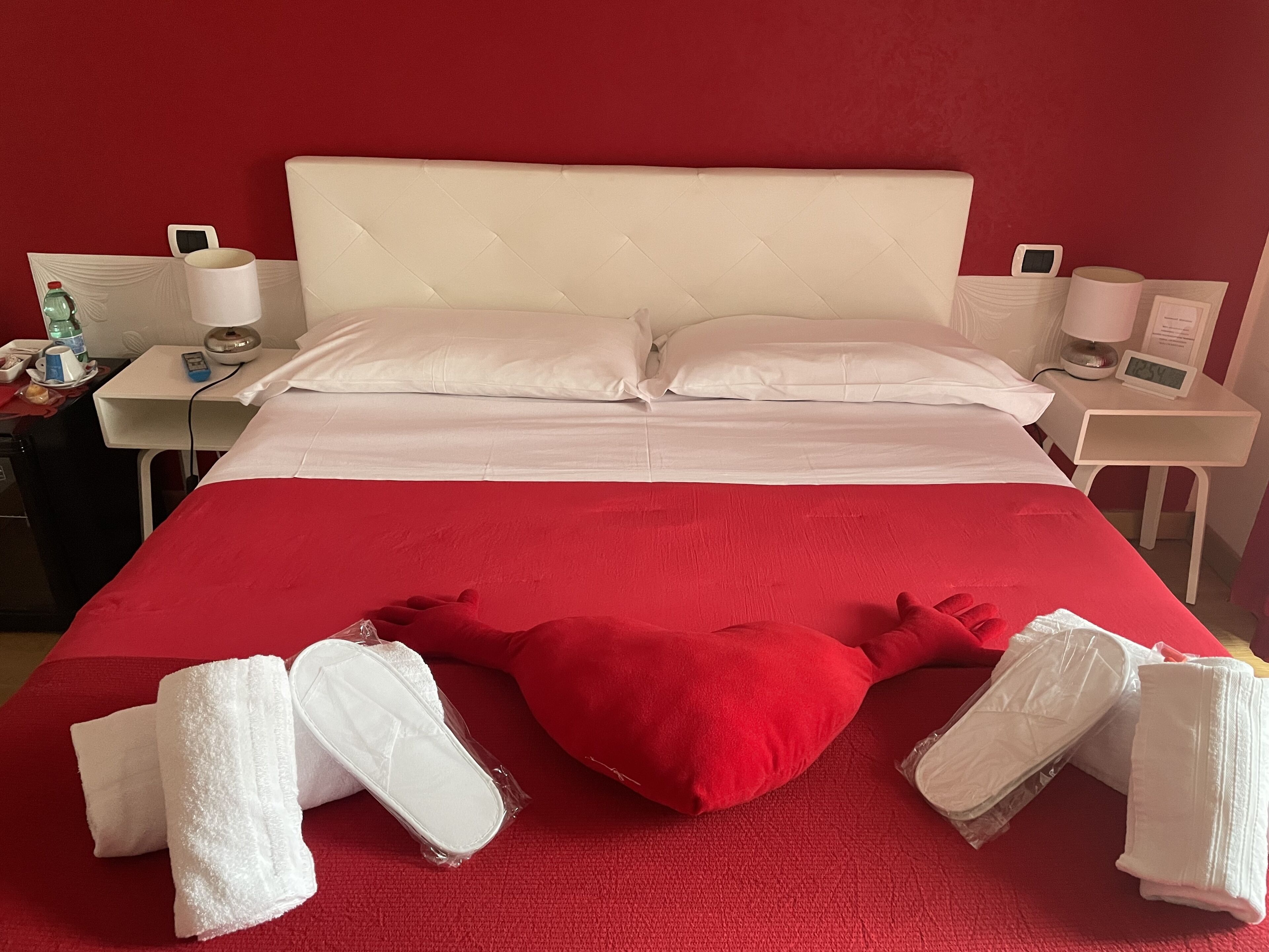 Een hotelkamer met een groot bed, witte lakens, een rood accentkussen, en een nachtkastje met een lamp en een digitale klok.
