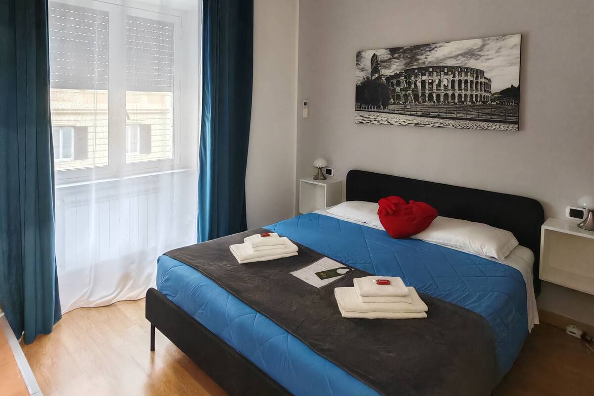 Habitación de hotel con cama, mesita de noche, lámpara de pared y ventana con cortinas.