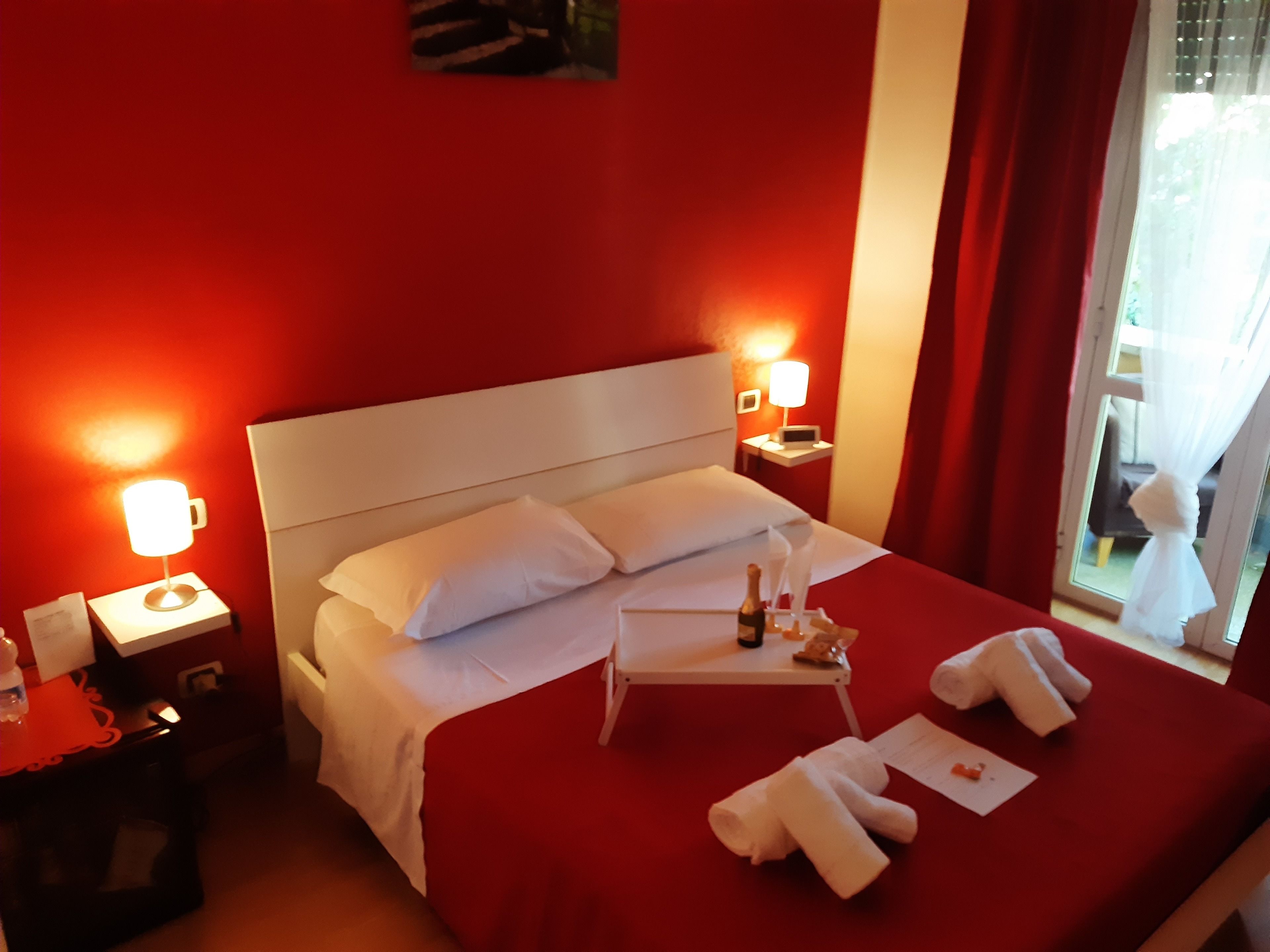 A hotel room with a red wall, white bed, and a nightstand with a lamp.