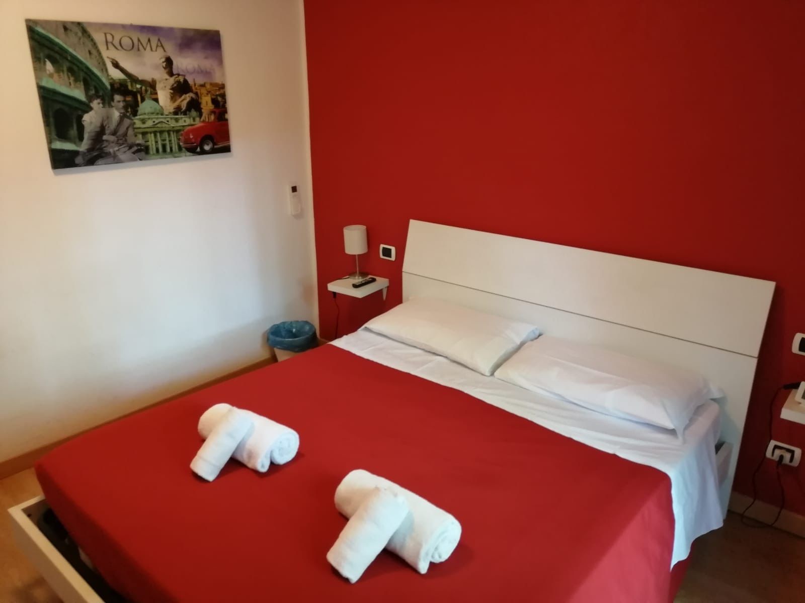 A hotel room with a red wall, a white bed, and a painting of Rome.