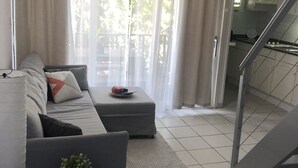 A modern living room with a grey sofa, a small table with a plant, and a kitchen area visible through a glass door. - Cozy one-bedroom & mezzanine apartment (Philipsburg)