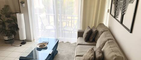 Een woonkamer met een glazen salontafel, een beige bank en uitzicht op een terras met stoelen.