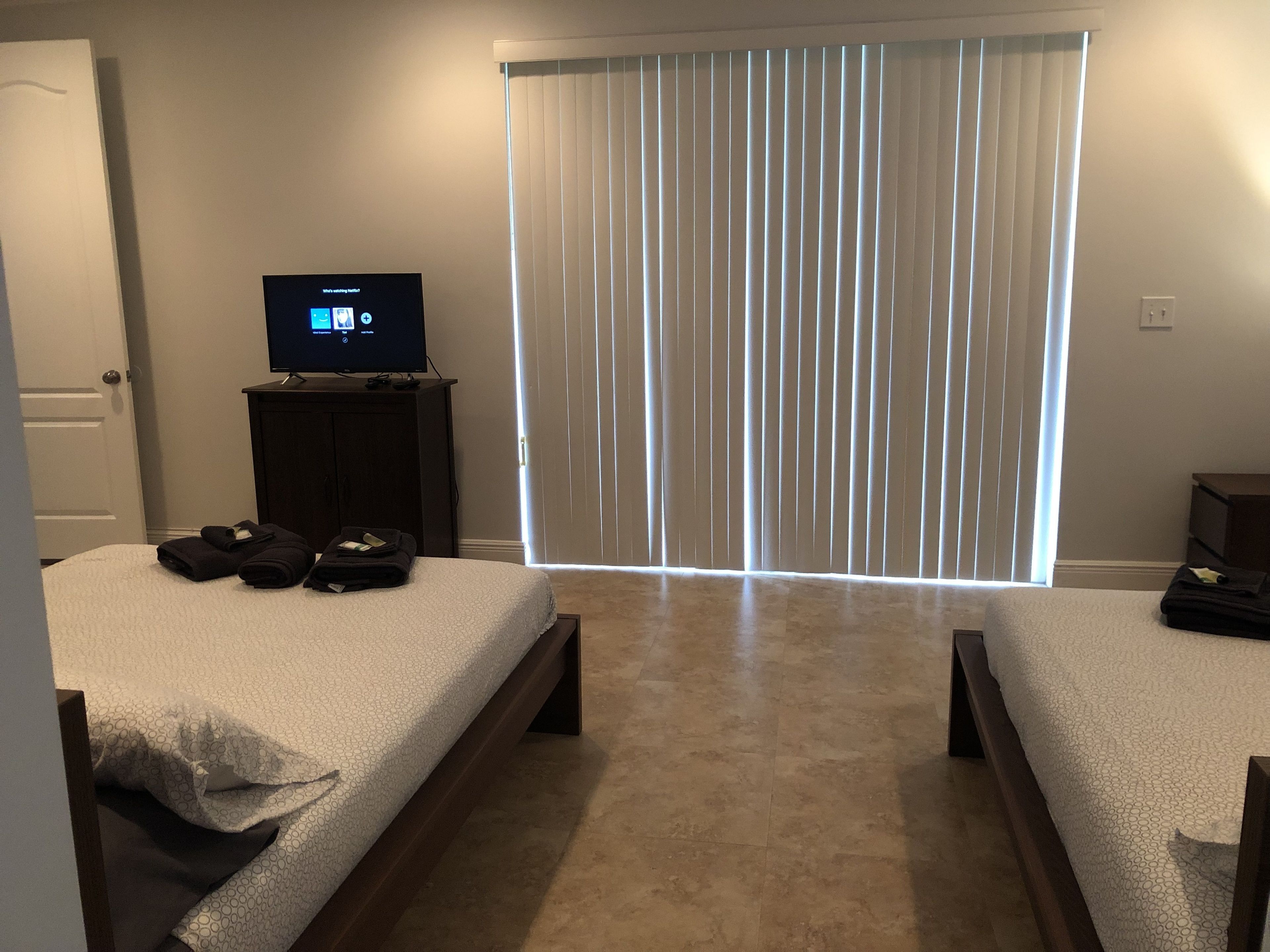 A hotel room with two beds, a TV on a stand, and a large window with blinds.