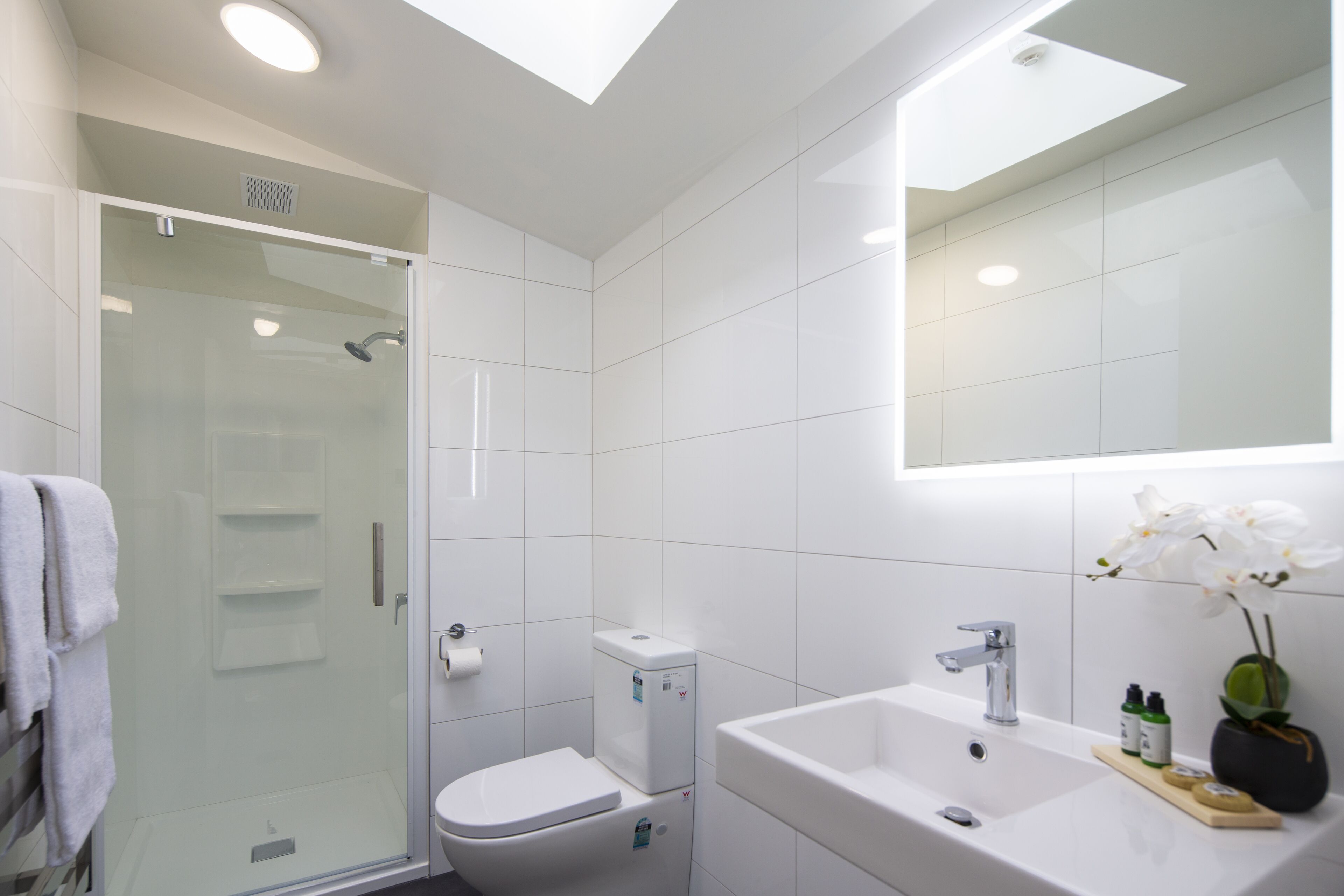 Ein modernes Badezimmer mit Duschkabine aus Glas, Toilette, Waschbecken und einem Spiegel darüber.