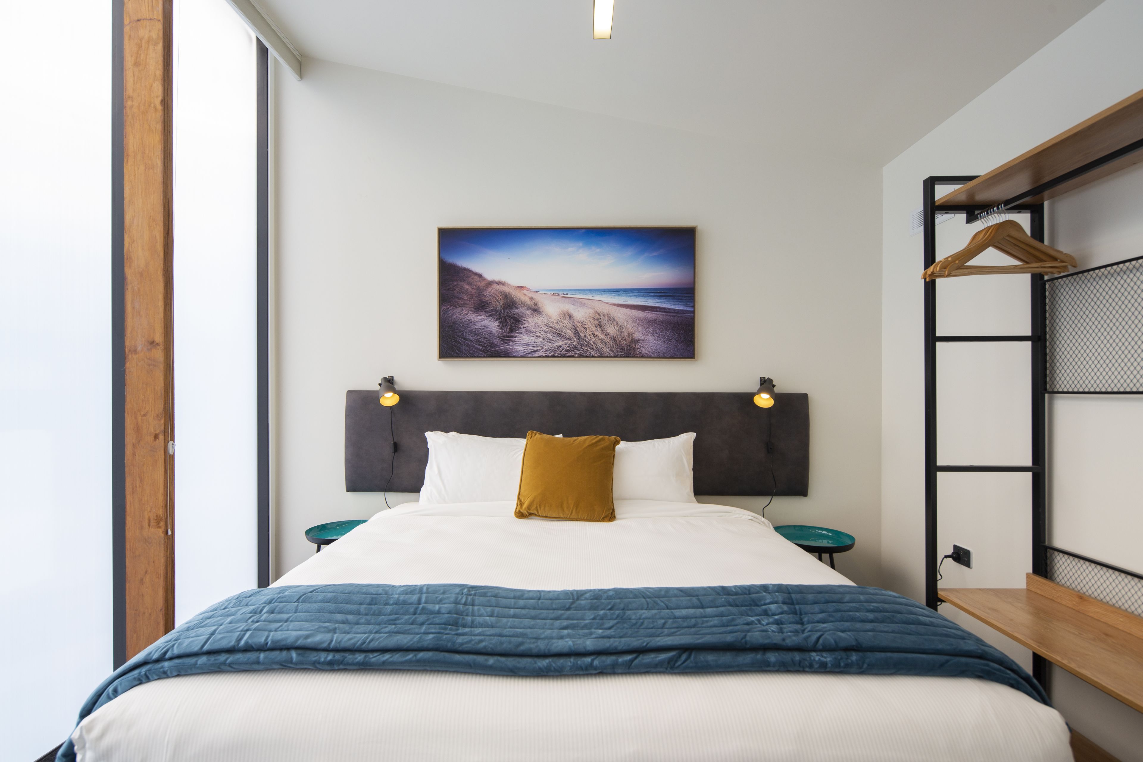 A neatly made bed with a blue quilt, a white pillow, and a mustard-colored pillow. A painting of a beach scene hangs above the bed. There is a small round table with a green top next to the bed.