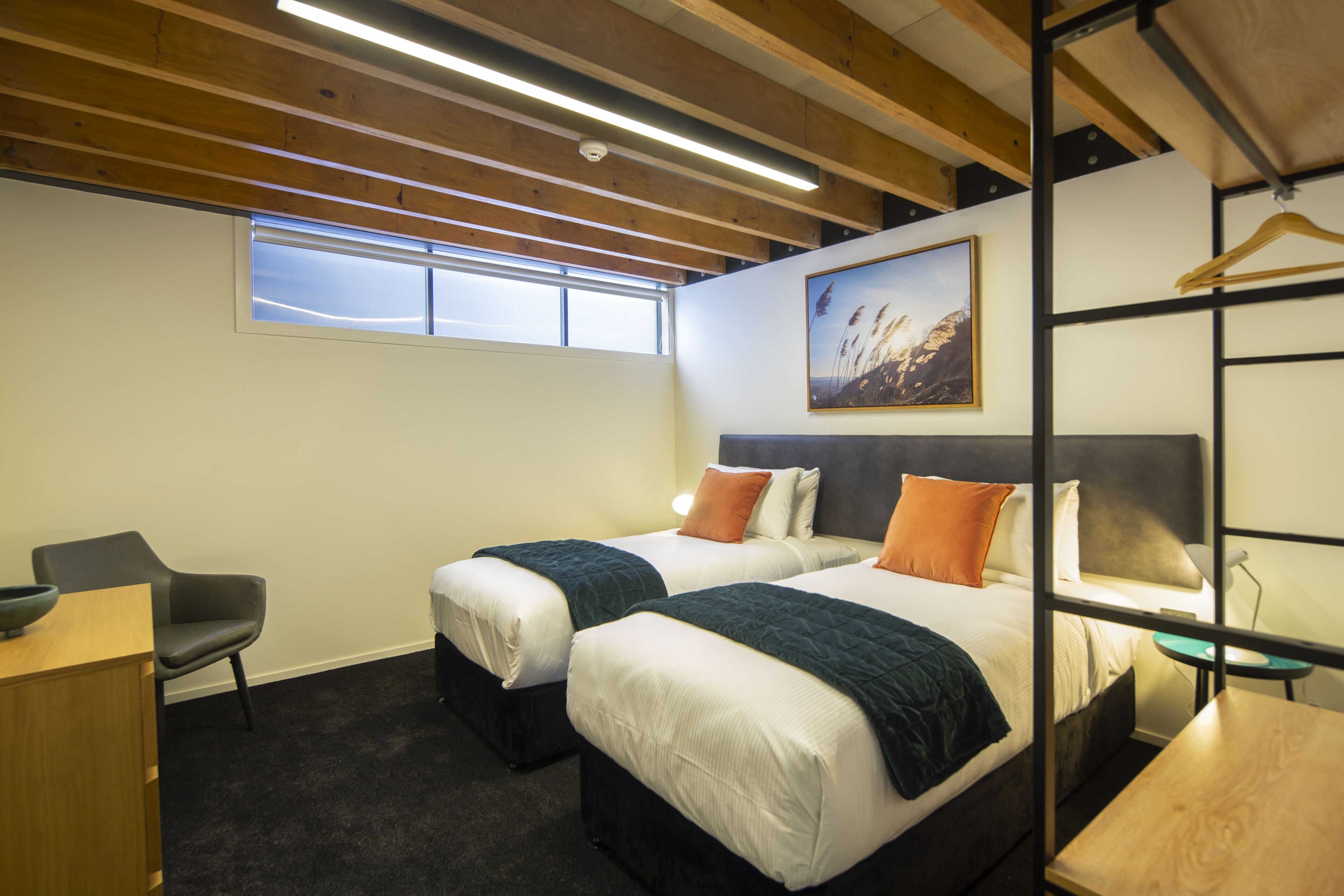 Quarto de hotel com duas camas, uma escrivaninha, uma cadeira, uma janela e um quadro na parede.