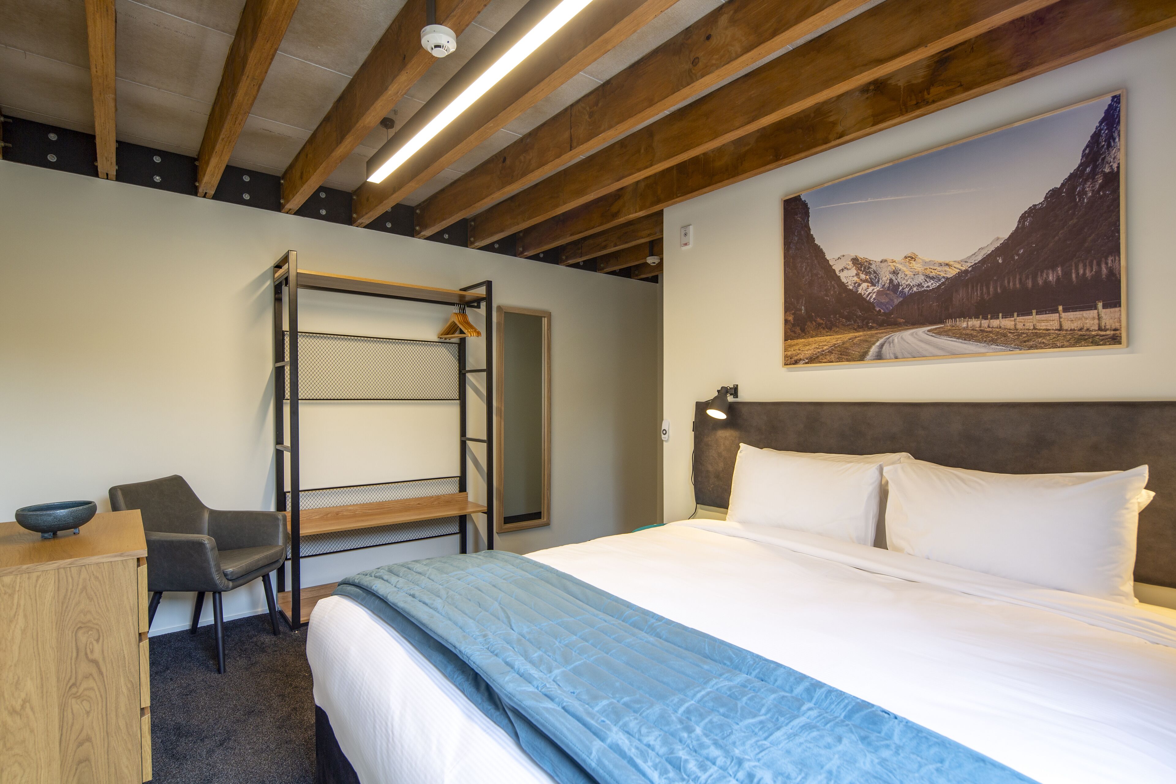 A hotel room with a bunk bed, a desk with a bowl, a chair, a bed with white linens, a headboard, and a mountain landscape painting on the wall.