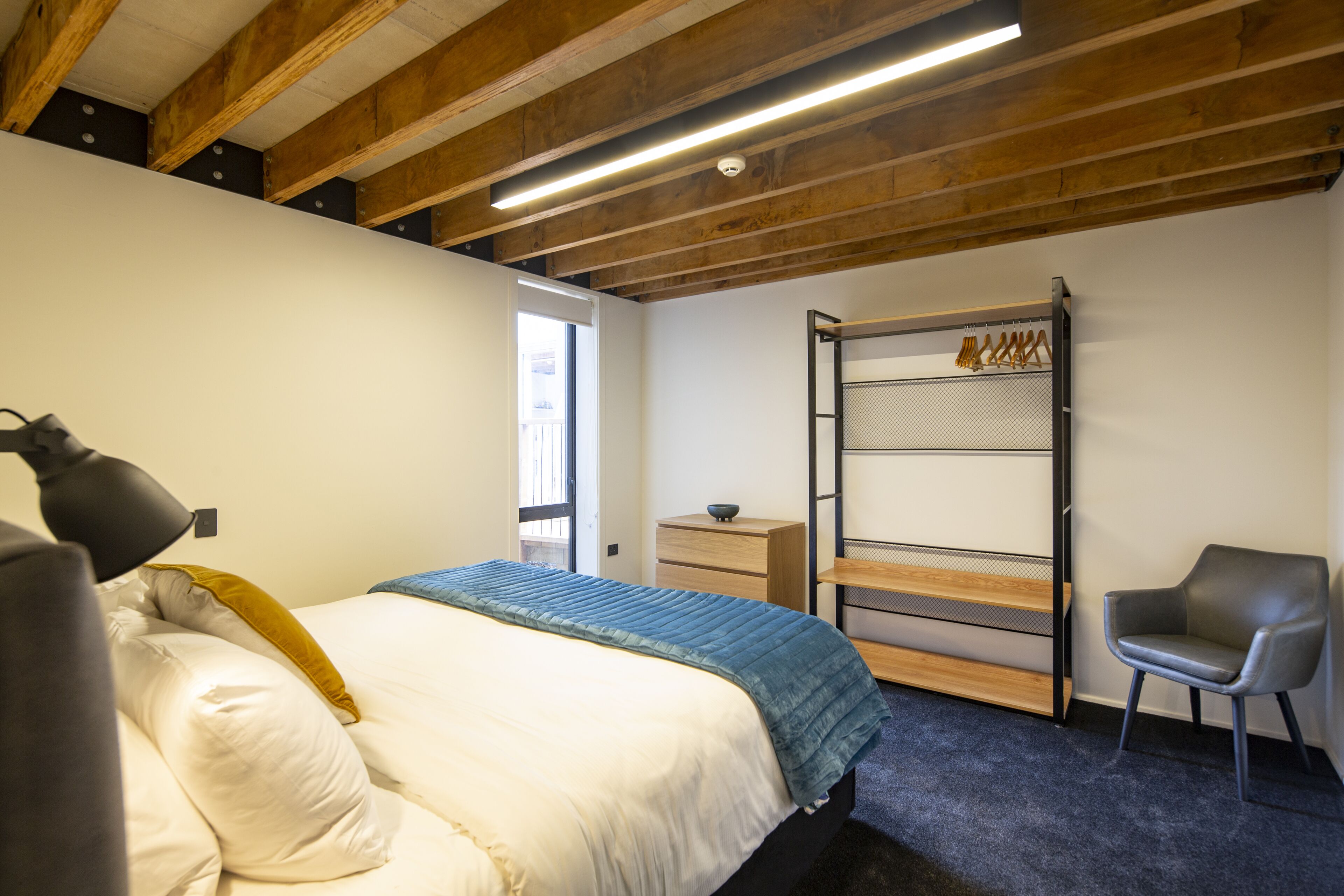 A bedroom with a bed, a chair, a desk, and a hanging rack.