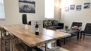 Ein geräumiger Wohnbereich mit Holztisch, Bänken, einer Couch und einem Couchtisch. An der Wand hängen gerahmte Bilder und auf dem Boden steht eine Stehlampe.