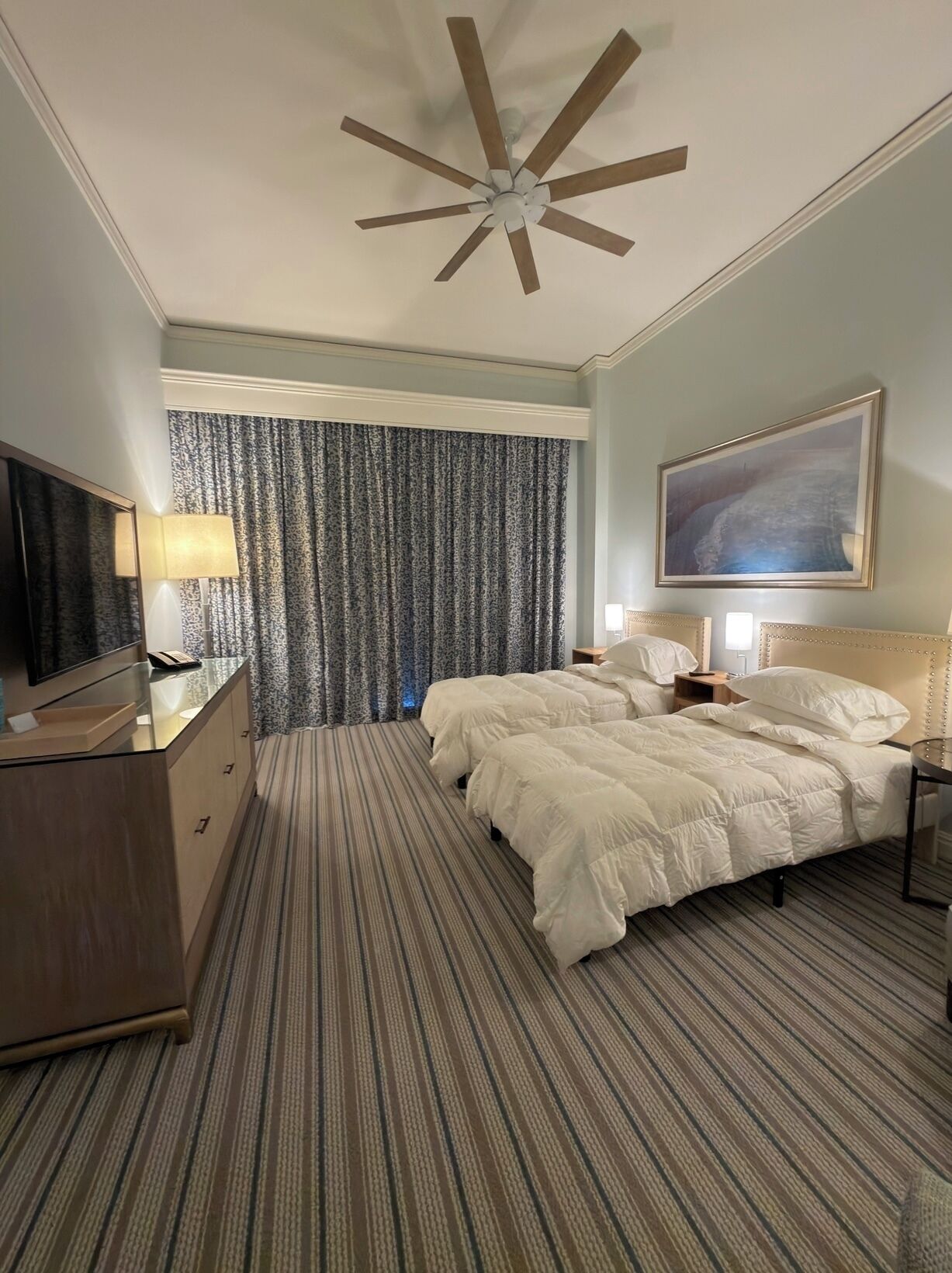A hotel room with a large bed, a ceiling fan, a TV, a desk, and a painting on the wall.