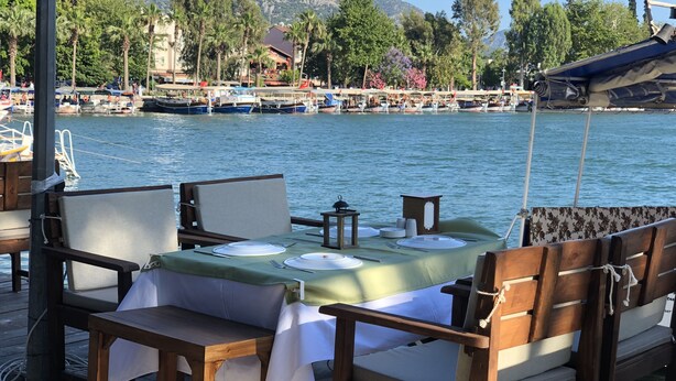 A dining area with a table set for two, overlooking a body of water with boats and a dock in the background. - Nehir Suit Hotel (Ortaca)