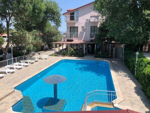 Una piscina con sillas de descanso y una sombrilla, rodeada de árboles y un edificio con balcones.