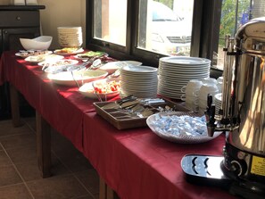 Una mesa de buffet con diversos alimentos, utensilios, una pila de platos y un dispensador de bebidas.