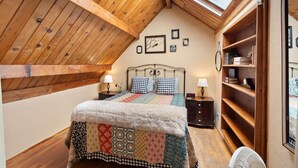 A bedroom with a sloped ceiling, a bed with a patterned quilt, bedside tables with lamps, a wooden bookshelf, and a skylight. - Night Owl - Murphys Vacation Rentals (Murphys)