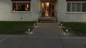 Un edificio de dos pisos con una entrada central, un balcón en el segundo nivel y un pequeño patio delantero con luces decorativas.