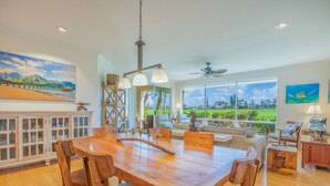 A spacious dining area with a wooden table and chairs, a ceiling fan, and large windows offering a view of a lush garden. - Relaxing Princeville Retreat • Golf Course + Sunset Views • A/C & Pool (Princeville)