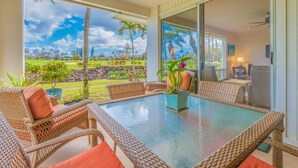 A sunlit patio with wicker furniture and a glass table, overlooking a lush garden and a golf course. - Relaxing Princeville Retreat • Golf Course + Sunset Views • A/C & Pool (Princeville)