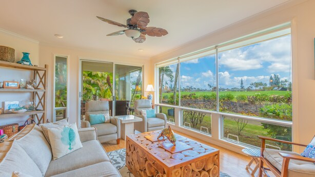 A spacious living room with a large window offering a view of a lush landscape, a ceiling fan, and a wooden coffee table. - Relaxing Princeville Retreat • Golf Course + Sunset Views • A/C & Pool (Princeville)