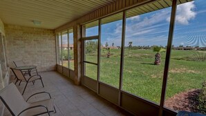 A covered patio with a brick wall, a glass door, and a view of a grassy field with palm trees.