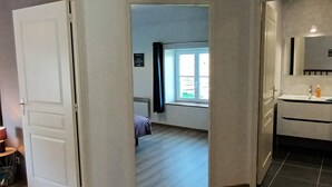 Ein Zimmer mit Bett, einem Fenster mit Vorhängen, einem kleinen Tisch und einem Badezimmer mit Spiegel und Waschbecken.