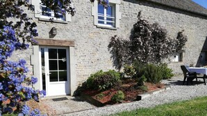 Ein Steinhaus mit einer weißen Tür, ein kleiner Garten mit Sträuchern und Blumen sowie ein Tisch mit Stühlen davor.