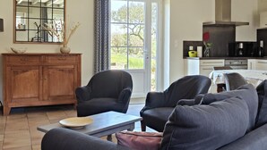 Ein Wohnzimmer mit einer Couch, zwei Sesseln, einem Couchtisch, einem Holzschrank und einer Küchenzeile mit Herd und Schränken.