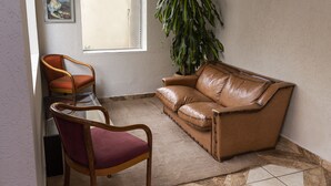 Una sala de estar con un sofá de cuero marrón, una silla de madera con un cojín rojo, una planta verde grande y un cuadro en la pared.