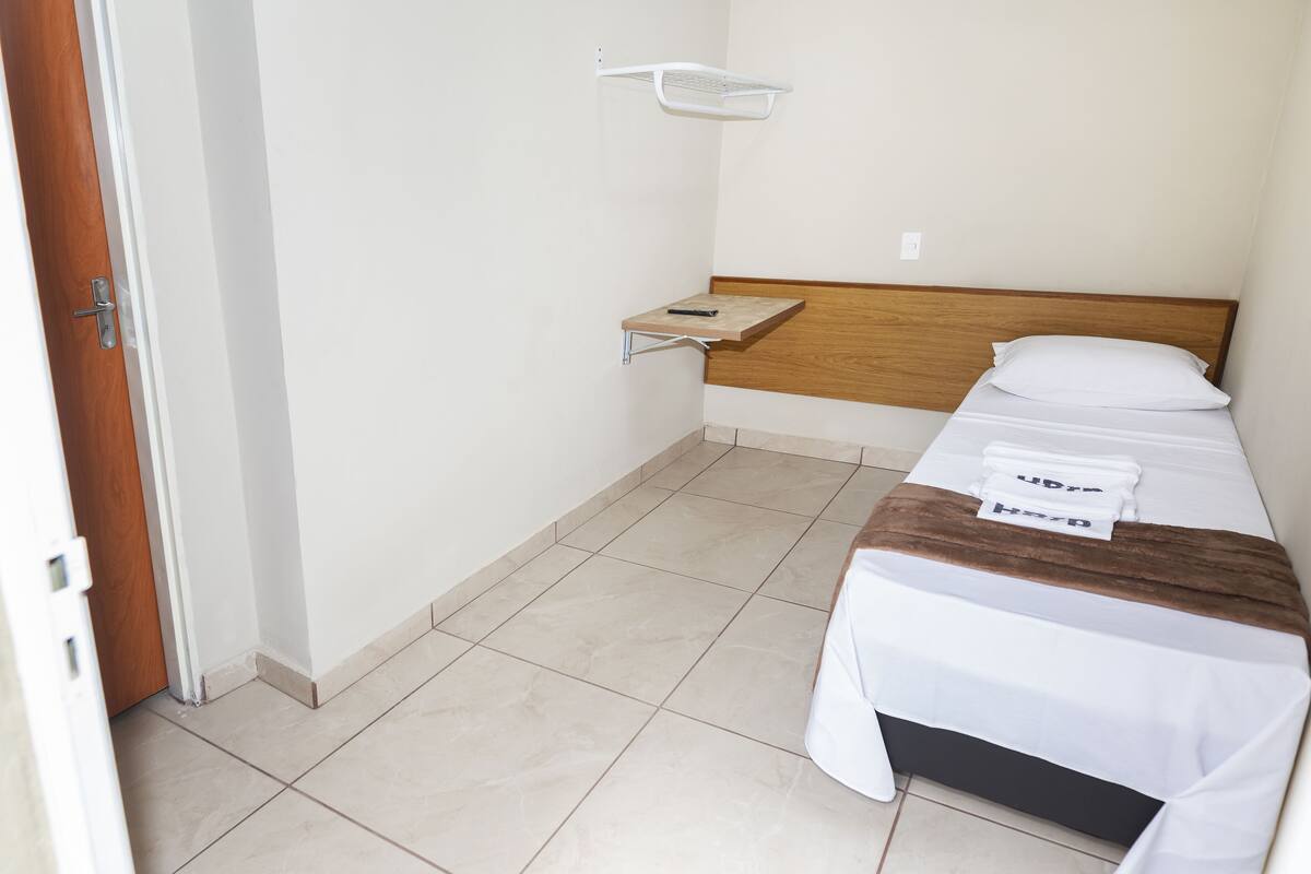 Ein kleines, sauberes Zimmer mit einem Bett, einem hölzernen Kopfteil, einem weißen Regal und einer weißen Tür.
