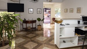 Una habitación amplia con piso de baldosas, un televisor de pantalla plana en la pared, una mesa de madera con sillas y un mostrador de recepción con una computadora y teléfono.