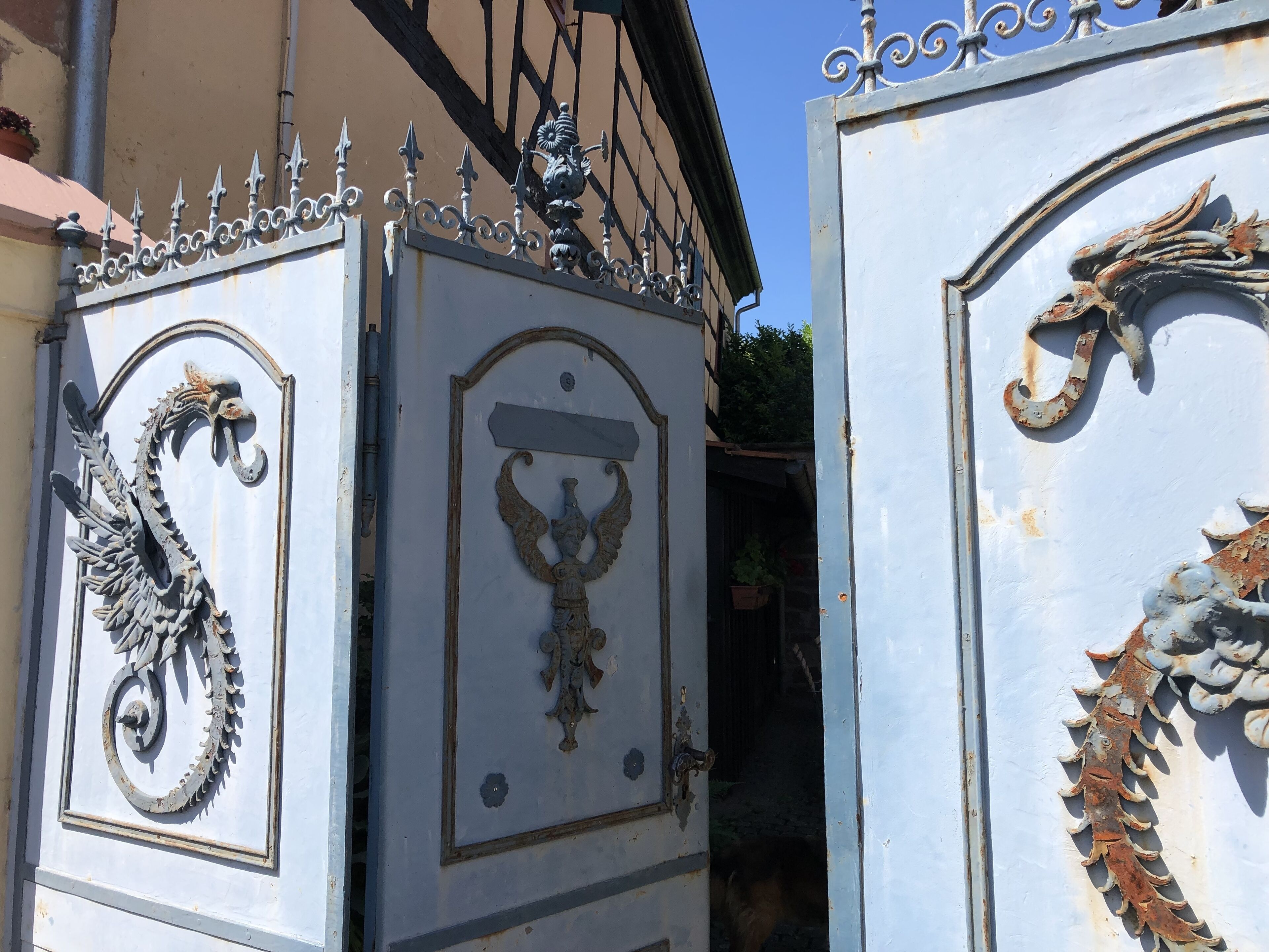Des grilles en métal ouvragé, ornées de motifs décoratifs, dont un dragon et un ange, mènent à l’entrée d’un bâtiment.