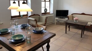 Comedor con una mesa de madera dispuesta para dos personas, un sofá, un televisor y un ventanal con cortinas.