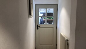 Un pasillo estrecho con una puerta blanca que tiene una ventana con persianas, un radiador a la derecha y una lámpara en el techo.