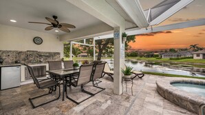 Un patio exterior con mesa y sillas, un ventilador de techo, zona de cocina y vistas a una piscina y al atardecer.