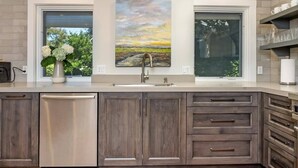 A modern kitchen with dark wood cabinets, a stainless steel dishwasher, and a large window with a view of greenery. - The Oxbow House - Designer 3BD/2BA Home (Napa)