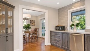 A modern kitchen with grey cabinets, a stainless steel dishwasher, and a window with a view of greenery. - The Oxbow House - Designer 3BD/2BA Home (Napa)