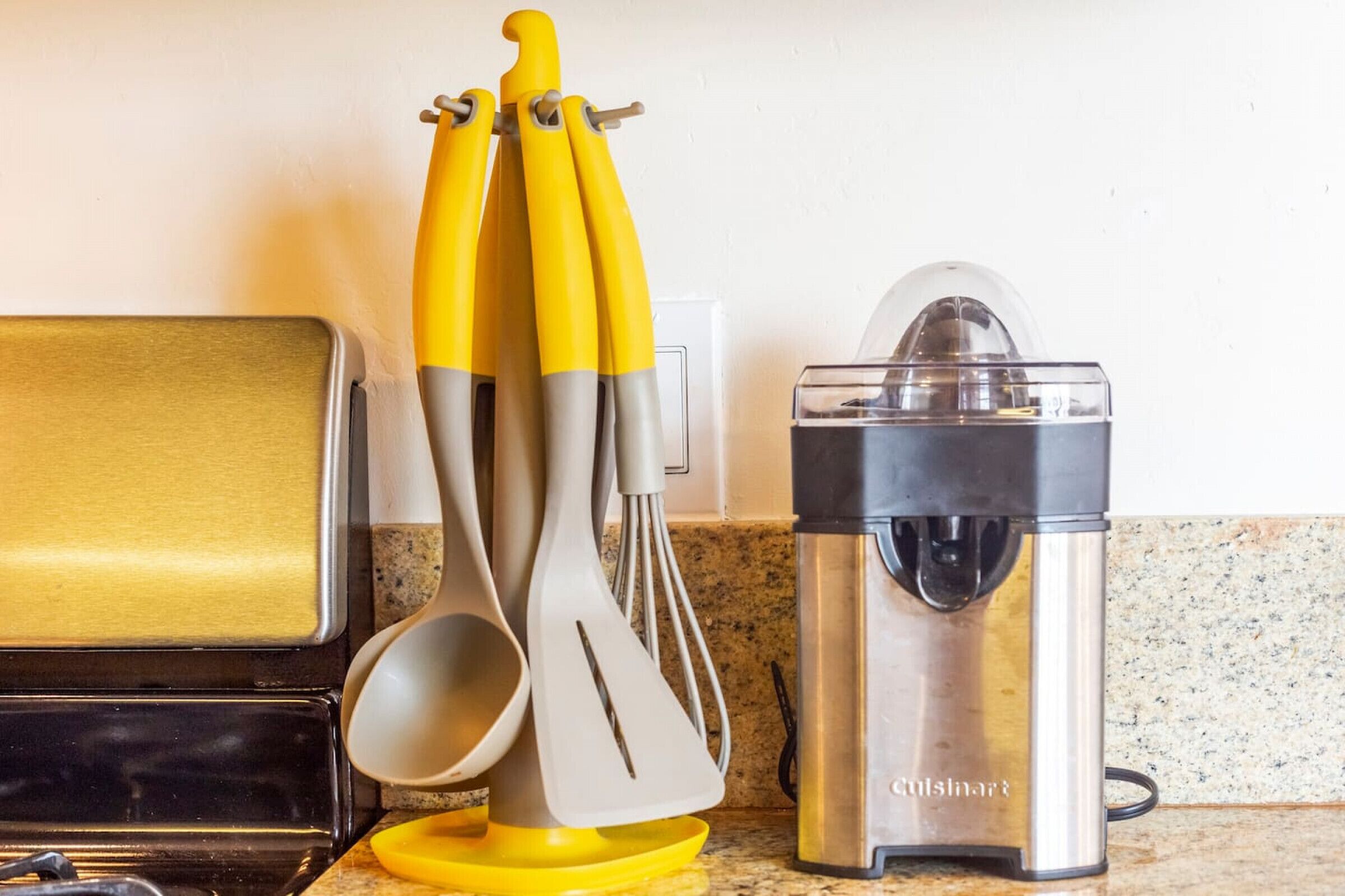 A kitchen counter with a food processor, a set of yellow and gray kitchen utensils, and a stainless steel appliance.