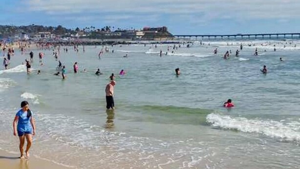 Una playa concurrida donde personas nadan y pasean por la orilla.