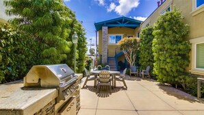 Un patio con una parrilla, mesa y sillas al aire libre, rodeado de follaje y un edificio con tejado azul.